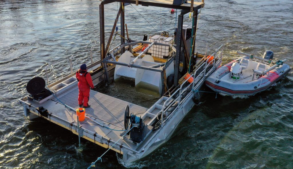 Crew members on a Waterotor test barge in a river, with a small boat alongside