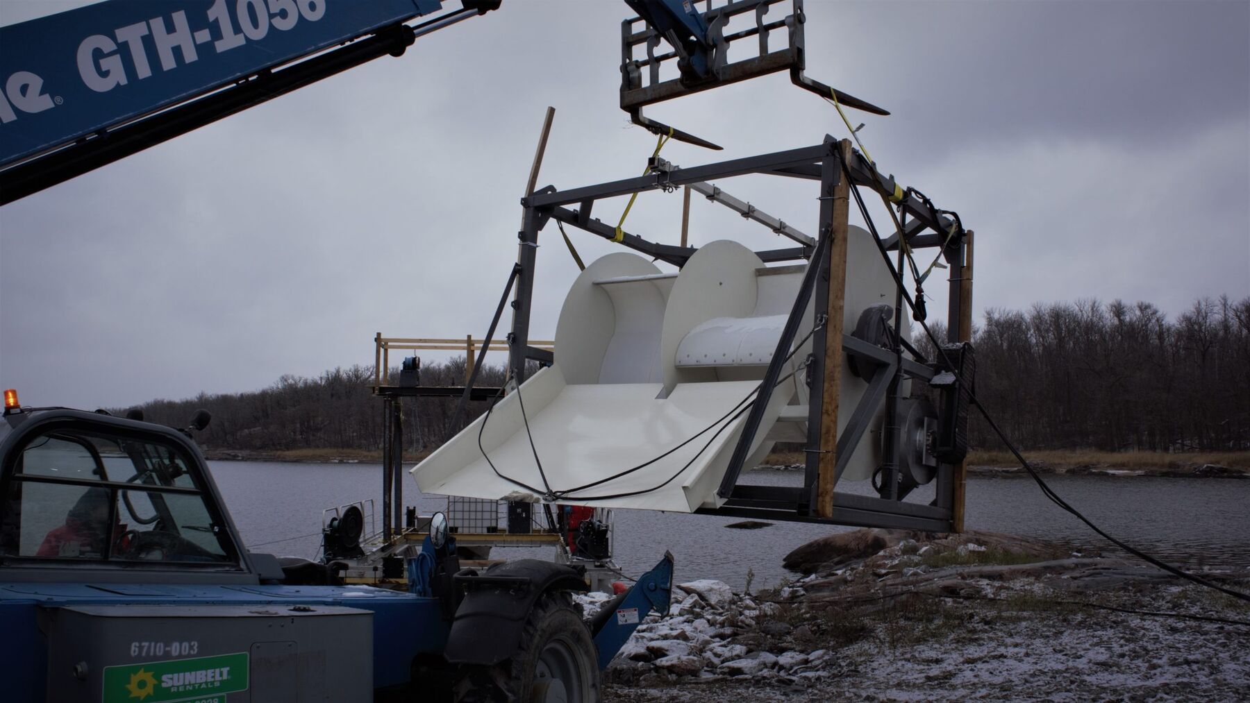 A crane lowers a white Waterotor on a test frame from the back of a truck into a cold northern river